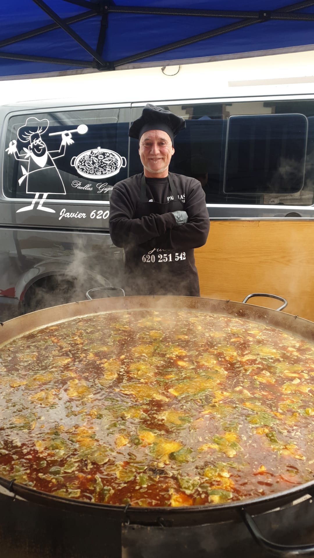 Chef Javier posando junto a una paella gigante de arroz con pollo, pato, ajos tiernos, habas y calabacín, cocinada con ingredientes frescos y técnicas tradicionales al aire libre.