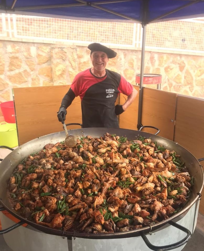 Chef sonriente cocinando una paella monumental con carne y verduras frescas en una gran paellera al aire libre, representando la tradición gastronómica de Guiral.