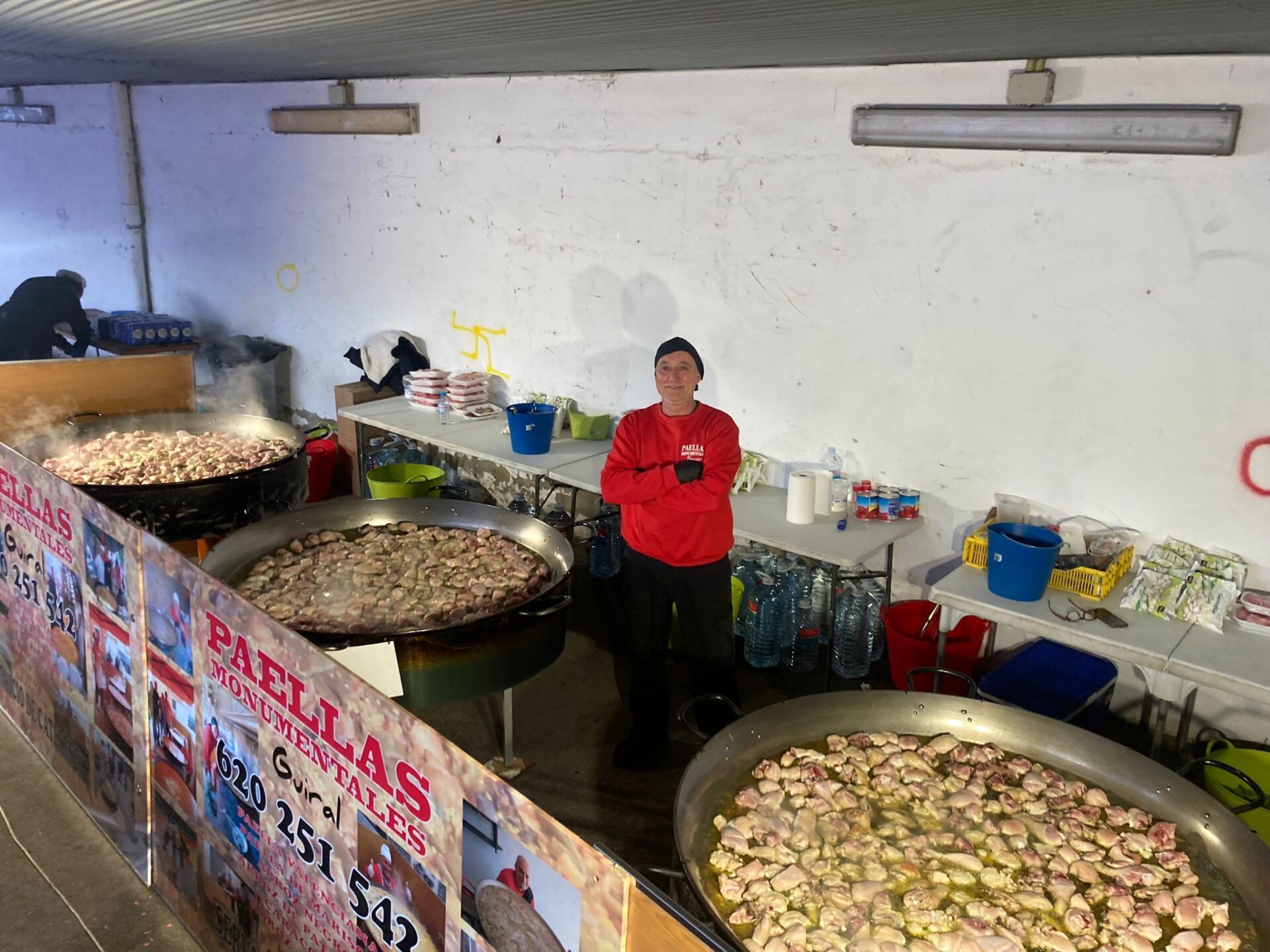 Hombre frente a paelleras gigantes en proceso de cocción de paellas monumentales con ingredientes frescos, parte de un evento culinario organizado por Guiral.