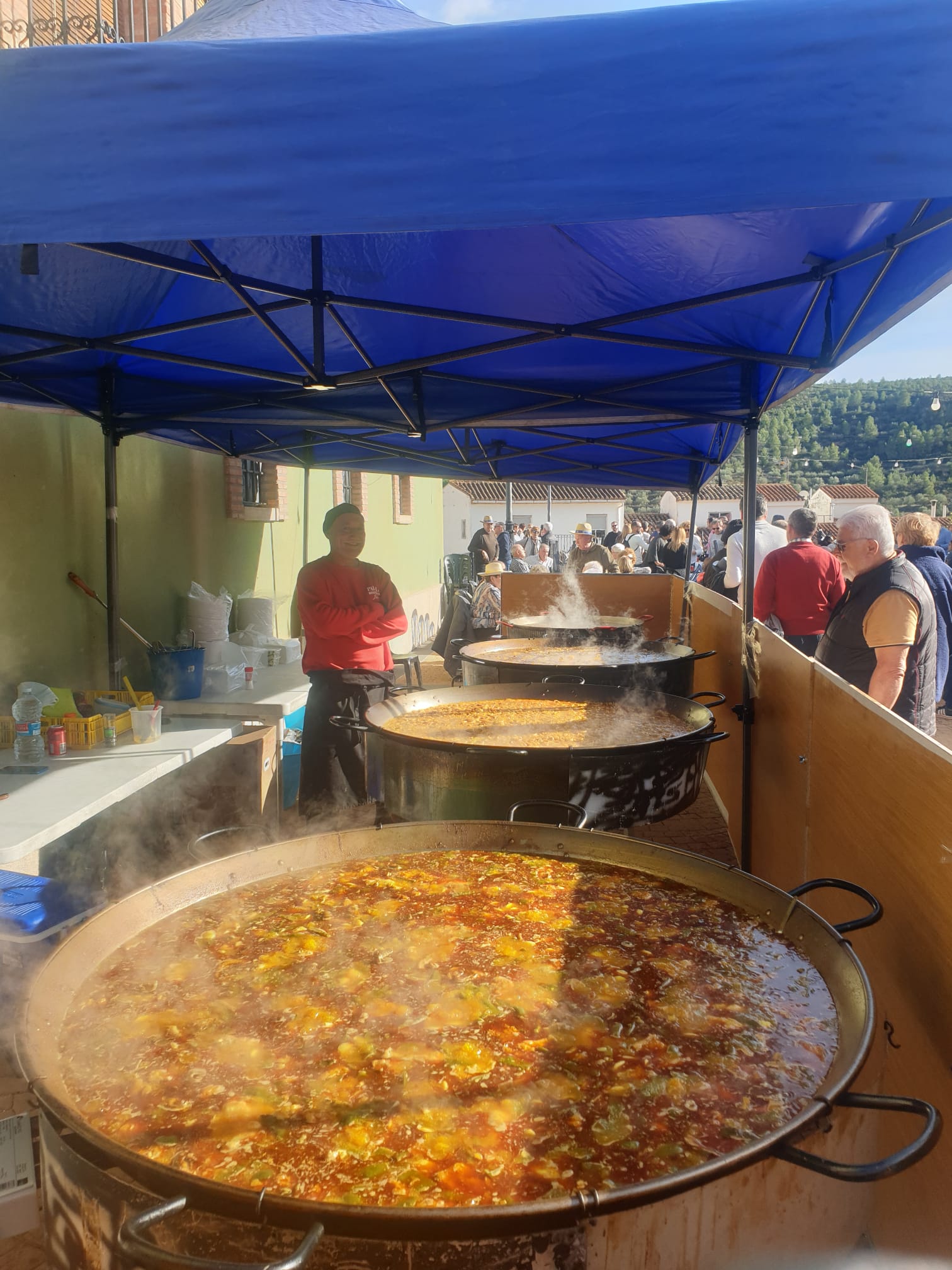 Imagen de un evento al aire libre con grandes paelleras llenas de coloridos ingredientes en plena cocción, organizada por Guiral, rodeada de asistentes disfrutando de la tradición.